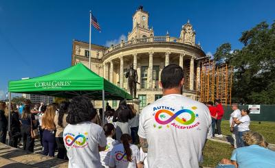 A crowd stands in front of city hall on a sunny day with blue sky. The main subjects in photo have their backs turned and are wearing Autism Acceptance shirts