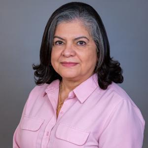 Monica Beltran poses for a headshot in a pink dress shirt in front of a gray background