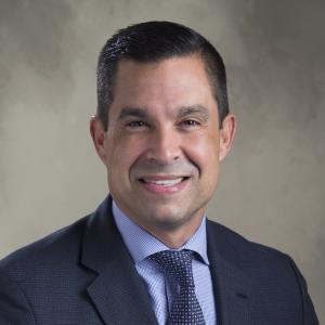 Vince Lago smiling in a suit with a beige background